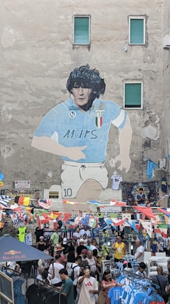 A large mural depicting a soccer player in a blue jersey is painted on a tall building. Below the mural, a bustling street market is set up with people shopping among numerous stalls. Brightly colored flags are strung across the scene, adding vibrance. Several people are browsing or purchasing merchandise, including sports jerseys.