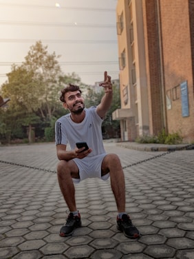 A man in a white sports outfit sits on a low chain while holding a smartphone. He is smiling and gesturing upward with his right hand. The background includes a brick building to the right, greenery, and a paved area with trees and power lines visible in the distance.