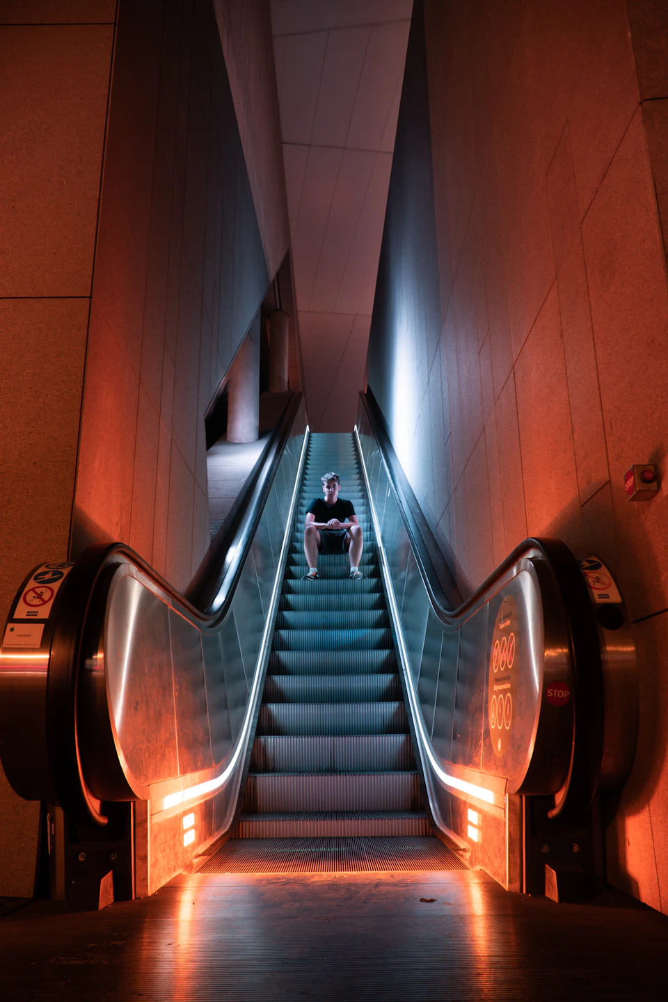 Man sits on escalator, gazing down at phone under warm orange lighting conditions
