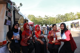 A group of five women, sitting together in an outdoor setting, are smiling and posing for the camera. They are wearing traditional attire with vibrant colors and are holding packaged food products. Tents and people in the background suggest a festival or market event.