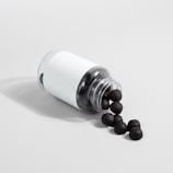 A transparent plastic bottle with a white label is lying on its side, spilling out several black sphere-shaped objects onto a light gray surface.