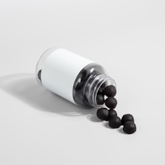 A transparent plastic bottle with a white label is lying on its side, spilling out several black sphere-shaped objects onto a light gray surface.