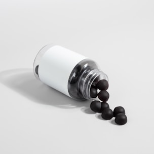 A transparent plastic bottle with a white label is lying on its side, spilling out several black sphere-shaped objects onto a light gray surface.