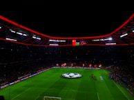 An illuminated football stadium with a large crowd and a group of people on the field, possibly preparing for a match. The roof has bright red lights and there are screens displaying content. A large graphic similar to a football competition emblem is visible on the field.
