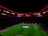 An illuminated football stadium with a large crowd and a group of people on the field, possibly preparing for a match. The roof has bright red lights and there are screens displaying content. A large graphic similar to a football competition emblem is visible on the field.
