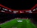 An illuminated football stadium with a large crowd and a group of people on the field, possibly preparing for a match. The roof has bright red lights and there are screens displaying content. A large graphic similar to a football competition emblem is visible on the field.