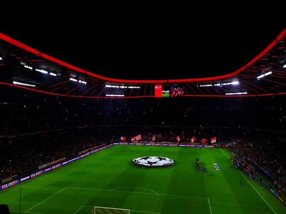 An illuminated football stadium with a large crowd and a group of people on the field, possibly preparing for a match. The roof has bright red lights and there are screens displaying content. A large graphic similar to a football competition emblem is visible on the field.
