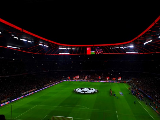 An illuminated football stadium with a large crowd and a group of people on the field, possibly preparing for a match. The roof has bright red lights and there are screens displaying content. A large graphic similar to a football competition emblem is visible on the field.