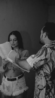 A black-and-white image featuring two people engaged in a conversation or discussion. The person on the left holds a few pages of paper and appears to be reading or discussing something with the other person, who is partly visible on the right side. The setting has minimalistic and natural lighting, creating a casual atmosphere.