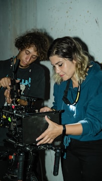 Two individuals are working together, focused and engaged in adjusting equipment on a film set. They are carefully handling a camera mounted on a tripod, with various buttons and settings being adjusted. Both appear to be in a dimly lit space, possibly a studio or location shoot.