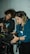Two individuals are working together, focused and engaged in adjusting equipment on a film set. They are carefully handling a camera mounted on a tripod, with various buttons and settings being adjusted. Both appear to be in a dimly lit space, possibly a studio or location shoot.