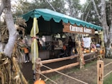 A festive outdoor booth with a green canopy labeled 'Carnival Games', surrounded by trees and decorated with cornstalks and ribbons. Several people, including children and adults, are engaged in activities, with bright orange pumpkins displayed around the booth.