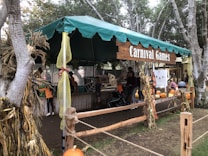 A festive outdoor booth with a green canopy labeled 'Carnival Games', surrounded by trees and decorated with cornstalks and ribbons. Several people, including children and adults, are engaged in activities, with bright orange pumpkins displayed around the booth.