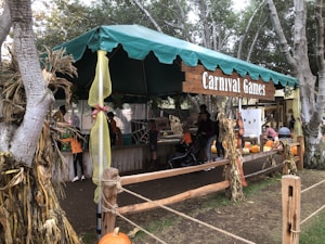 A festive outdoor booth with a green canopy labeled 'Carnival Games', surrounded by trees and decorated with cornstalks and ribbons. Several people, including children and adults, are engaged in activities, with bright orange pumpkins displayed around the booth.