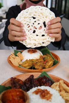 A person holds a large, circular, intricately woven rice cracker in both hands. In front, there are two wooden plates with servings of rice topped with fried shallots, accompanied by side dishes such as spicy chicken, sambal, and crispy crackers, all placed on banana leaves.
