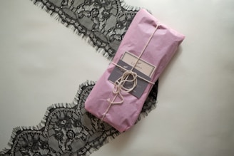 A neatly wrapped package in pink paper is tied with twine, placed on top of an elegant black lace fabric. The package includes a label that suggests a sophisticated product presentation.