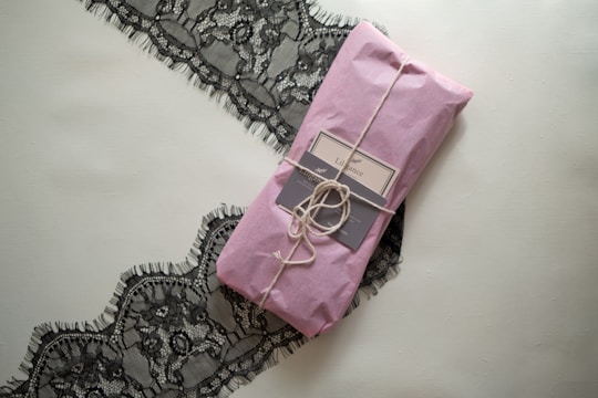 A neatly wrapped package in pink paper is tied with twine, placed on top of an elegant black lace fabric. The package includes a label that suggests a sophisticated product presentation.