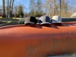 Three baseball caps in black, dark gray, and light gray are lined up on an orange metal surface outdoors. The background features blurred trees and a clear blue sky, suggesting a sunny day in a park or natural setting.