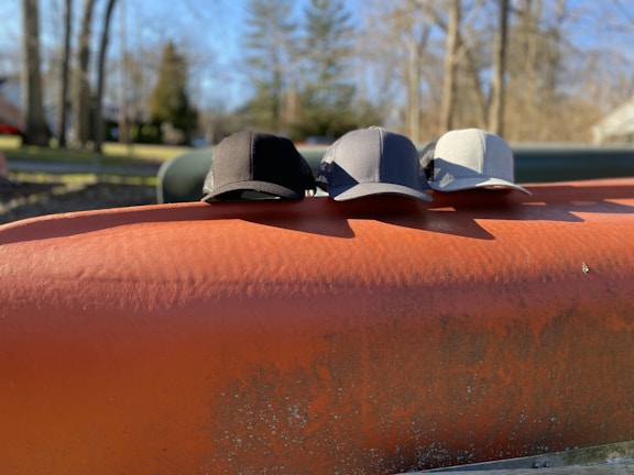 Three baseball caps in black, dark gray, and light gray are lined up on an orange metal surface outdoors. The background features blurred trees and a clear blue sky, suggesting a sunny day in a park or natural setting.