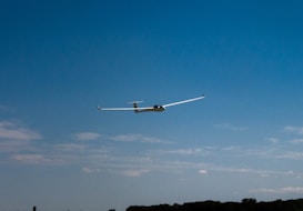 A glider aircraft is soaring through a clear blue sky. The landscape below features a horizon line with a few scattered clouds.
