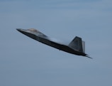A modern fighter jet is captured in flight, angled upward against a backdrop of a clear blue sky. The aircraft appears aerodynamic and sleek, emitting vapor trails from its wings, which suggests high-speed movement.