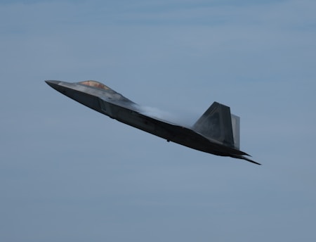 A modern fighter jet is captured in flight, angled upward against a backdrop of a clear blue sky. The aircraft appears aerodynamic and sleek, emitting vapor trails from its wings, which suggests high-speed movement.
