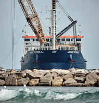 A large ship with construction equipment, including a crane arm, positioned at the port. The vessel has a dark blue hull and white superstructure with a visible name on the side. The foreground features a rocky breakwater and gentle waves on the water surface.