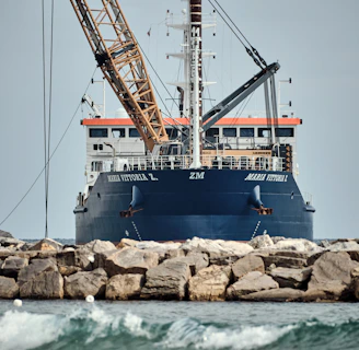 A large ship with construction equipment, including a crane arm, positioned at the port. The vessel has a dark blue hull and white superstructure with a visible name on the side. The foreground features a rocky breakwater and gentle waves on the water surface.
