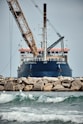 A large ship with construction equipment, including a crane arm, positioned at the port. The vessel has a dark blue hull and white superstructure with a visible name on the side. The foreground features a rocky breakwater and gentle waves on the water surface.