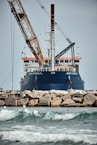 A large ship with construction equipment, including a crane arm, positioned at the port. The vessel has a dark blue hull and white superstructure with a visible name on the side. The foreground features a rocky breakwater and gentle waves on the water surface.