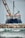 A large ship with construction equipment, including a crane arm, positioned at the port. The vessel has a dark blue hull and white superstructure with a visible name on the side. The foreground features a rocky breakwater and gentle waves on the water surface.