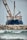 A large ship with construction equipment, including a crane arm, positioned at the port. The vessel has a dark blue hull and white superstructure with a visible name on the side. The foreground features a rocky breakwater and gentle waves on the water surface.