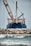 A large ship with construction equipment, including a crane arm, positioned at the port. The vessel has a dark blue hull and white superstructure with a visible name on the side. The foreground features a rocky breakwater and gentle waves on the water surface.