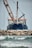 A large ship with construction equipment, including a crane arm, positioned at the port. The vessel has a dark blue hull and white superstructure with a visible name on the side. The foreground features a rocky breakwater and gentle waves on the water surface.