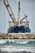A large ship with construction equipment, including a crane arm, positioned at the port. The vessel has a dark blue hull and white superstructure with a visible name on the side. The foreground features a rocky breakwater and gentle waves on the water surface.