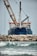 A large ship with construction equipment, including a crane arm, positioned at the port. The vessel has a dark blue hull and white superstructure with a visible name on the side. The foreground features a rocky breakwater and gentle waves on the water surface.