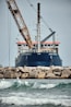 A large ship with construction equipment, including a crane arm, positioned at the port. The vessel has a dark blue hull and white superstructure with a visible name on the side. The foreground features a rocky breakwater and gentle waves on the water surface.
