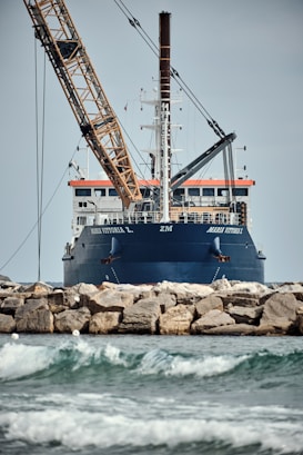A large ship with construction equipment, including a crane arm, positioned at the port. The vessel has a dark blue hull and white superstructure with a visible name on the side. The foreground features a rocky breakwater and gentle waves on the water surface.