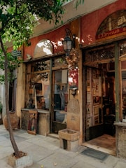 A quaint and rustic exterior of a shop or cafe, adorned with vintage decorations. The storefront features wooden doors and windows framed by an assortment of artworks and decor. A small, circular basin with intricate carvings is positioned near the entrance. There is a tree providing shade and a traditional lantern.