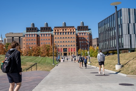 A group of people walk along a wide concrete pathway leading to a large, modern academic building with a distinctive architectural style. The building is surrounded by green grass and trees with autumn foliage. On either side of the path, there are metal lamp posts and modern office buildings.
