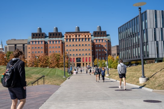 A group of people walk along a wide concrete pathway leading to a large, modern academic building with a distinctive architectural style. The building is surrounded by green grass and trees with autumn foliage. On either side of the path, there are metal lamp posts and modern office buildings.