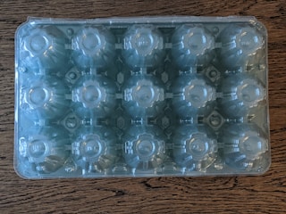 A clear plastic egg carton is placed on a textured wooden surface, displaying slots for 18 eggs.