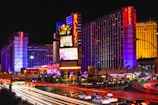 Brightly lit cityscape at night featuring a large hotel with neon signs displaying the name Bally's. The structure is illuminated with purple and blue lights, and colorful streaks from car lights create a vibrant atmosphere. Additional signage advertises various attractions and businesses.