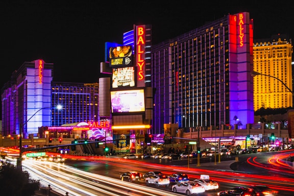 Brightly lit cityscape at night featuring a large hotel with neon signs displaying the name Bally's. The structure is illuminated with purple and blue lights, and colorful streaks from car lights create a vibrant atmosphere. Additional signage advertises various attractions and businesses.