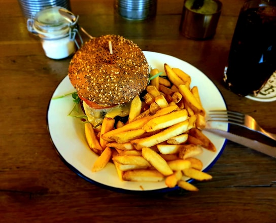 A sesame seed bun hamburger is served on a white plate alongside a generous portion of golden, crispy French fries. The burger is stacked with ingredients like lettuce and a slice of tomato, and topped with melted cheese. A soda in a glass sits in the background on a wooden table, along with a small jar and other condiments.