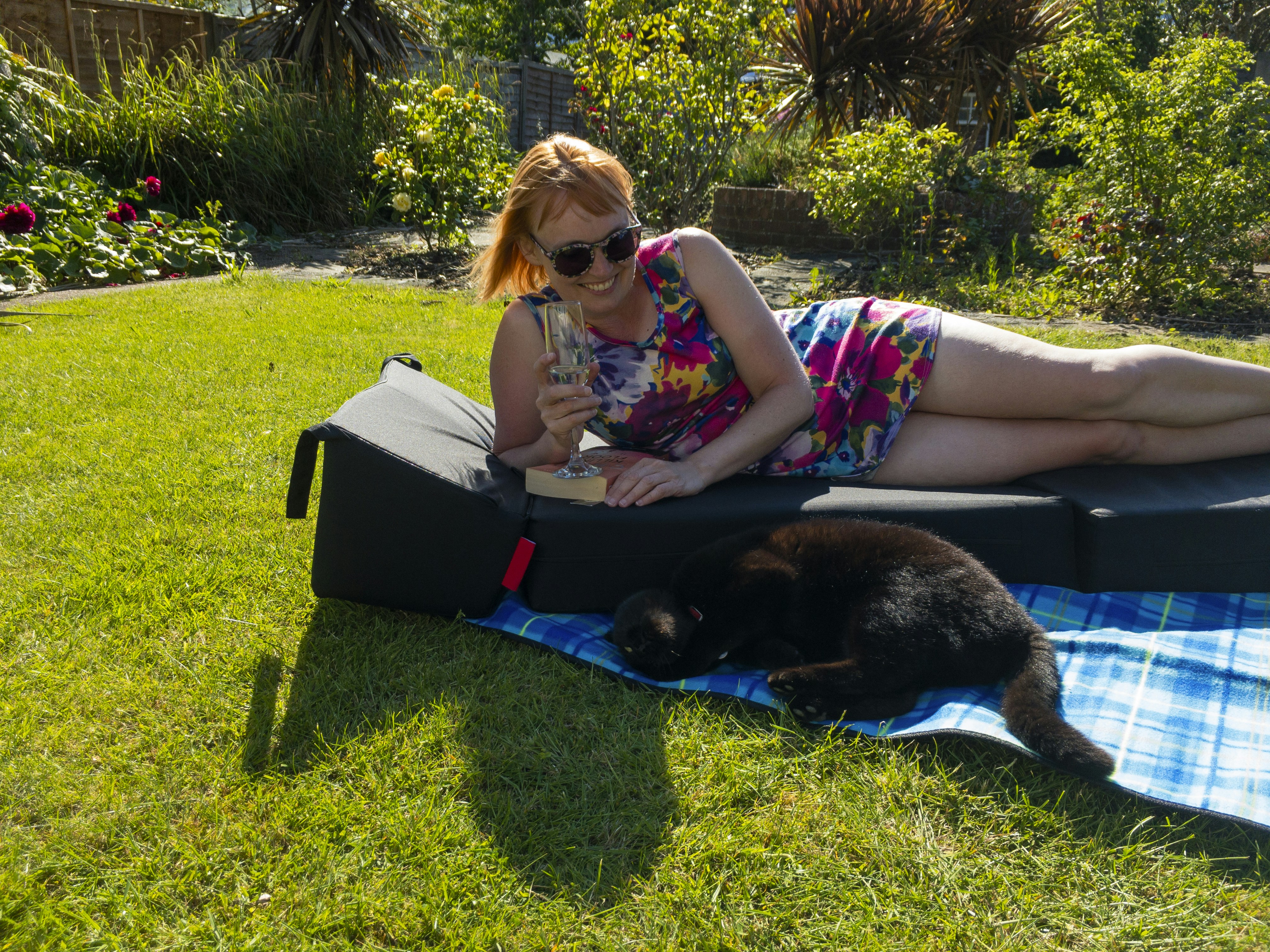 Smiling woman wearing sunglasses in a short, colourful summer dress lying in the sun on a padded lounger and a blue picnic blanket in the garden holding a glass of champagne with her cat lying next to her