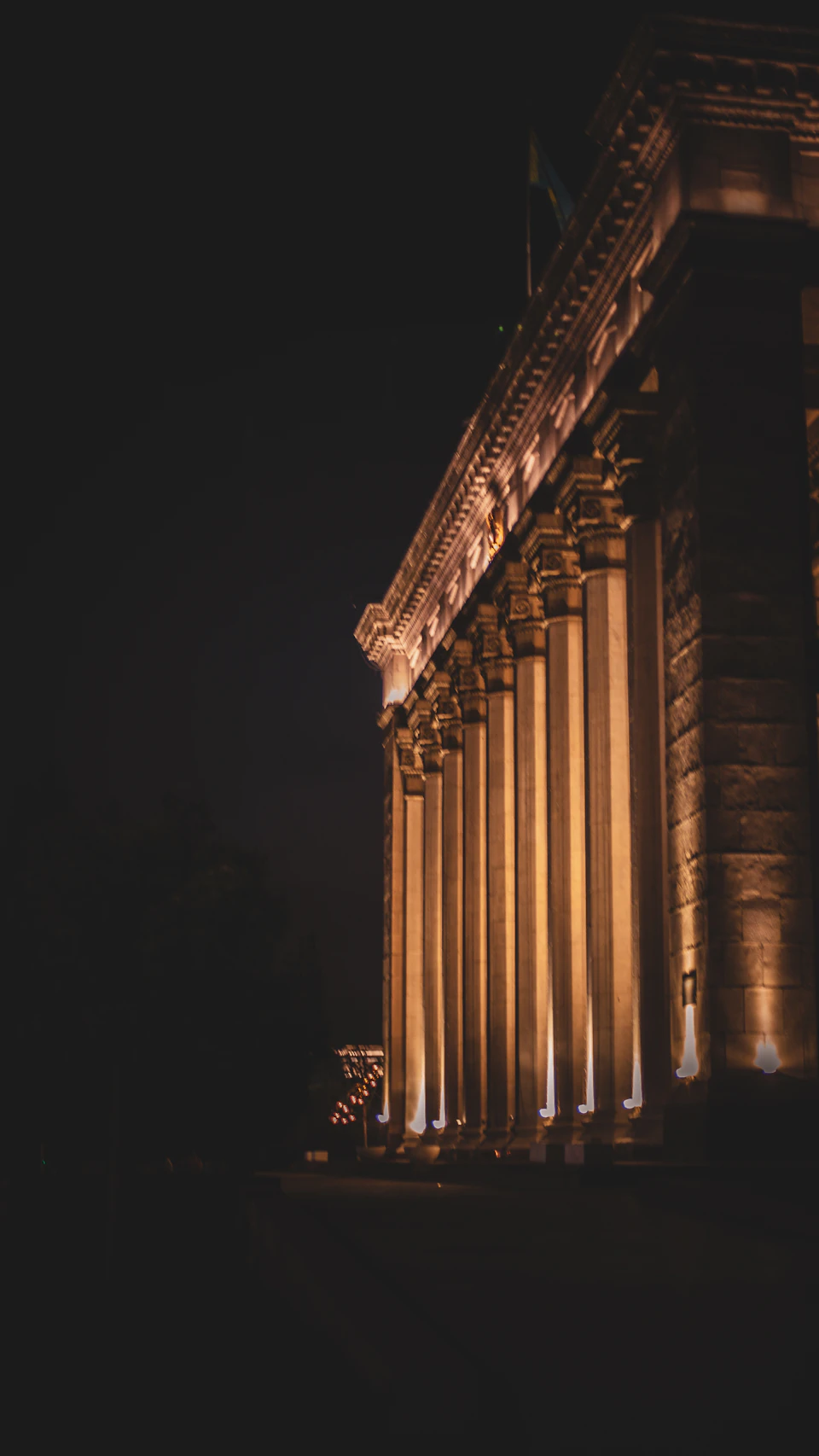 The image depicts a grand, ornate building with numerous columns and arches, bathed in warm yellow light against a dark night sky. The building's facade is illuminated from below, creating a striking contrast between the illuminated structure and its surroundings. In the foreground, there are trees that add depth to the scene, providing a sense of scale and perspective.

The image does not contain any discernible text or countable objects, but it captures the architectural beauty of the building in a serene nighttime setting.