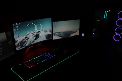 A dark room illuminated by two computer monitors displaying desktop backgrounds with mountain images. A gaming setup features a keyboard with RGB lighting and a tower with LED fans glowing in blue and purple.