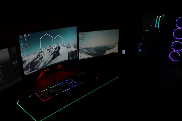 A dark room illuminated by two computer monitors displaying desktop backgrounds with mountain images. A gaming setup features a keyboard with RGB lighting and a tower with LED fans glowing in blue and purple.