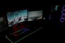 A dark room illuminated by two computer monitors displaying desktop backgrounds with mountain images. A gaming setup features a keyboard with RGB lighting and a tower with LED fans glowing in blue and purple.